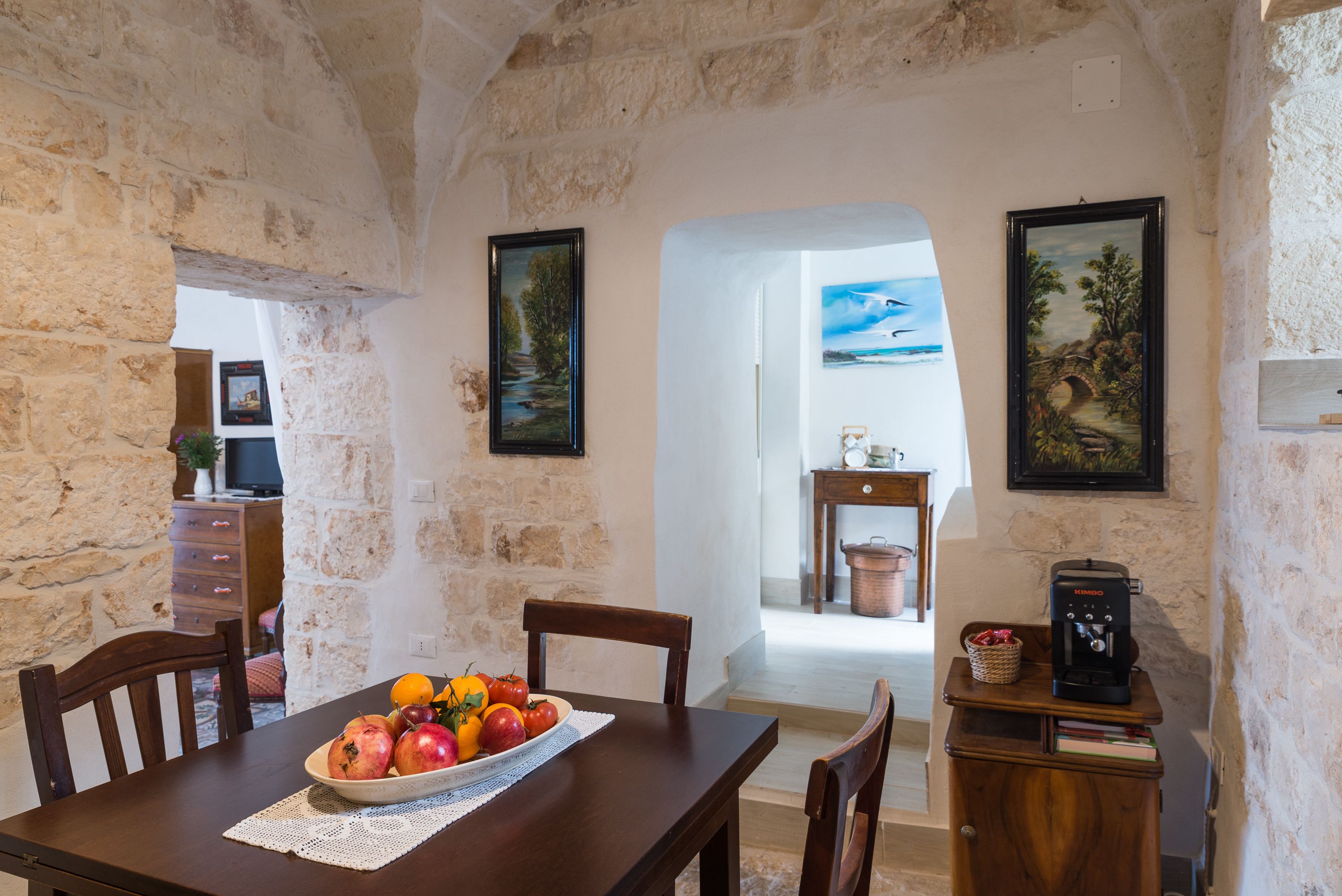 A rustic dining area with a wooden table, chairs, and a bowl of fruit. There are stone walls and a painting on the wall.