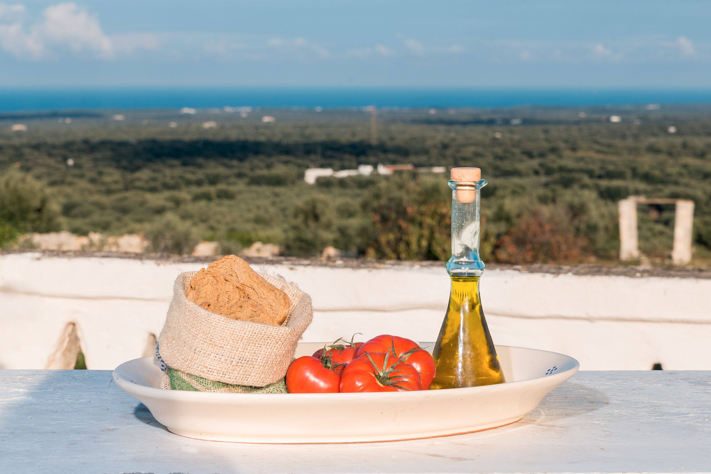 A table with a plate containing bread, tomatoes, and a bottle of olive oil, set against a scenic backdrop of a coastal landscape.