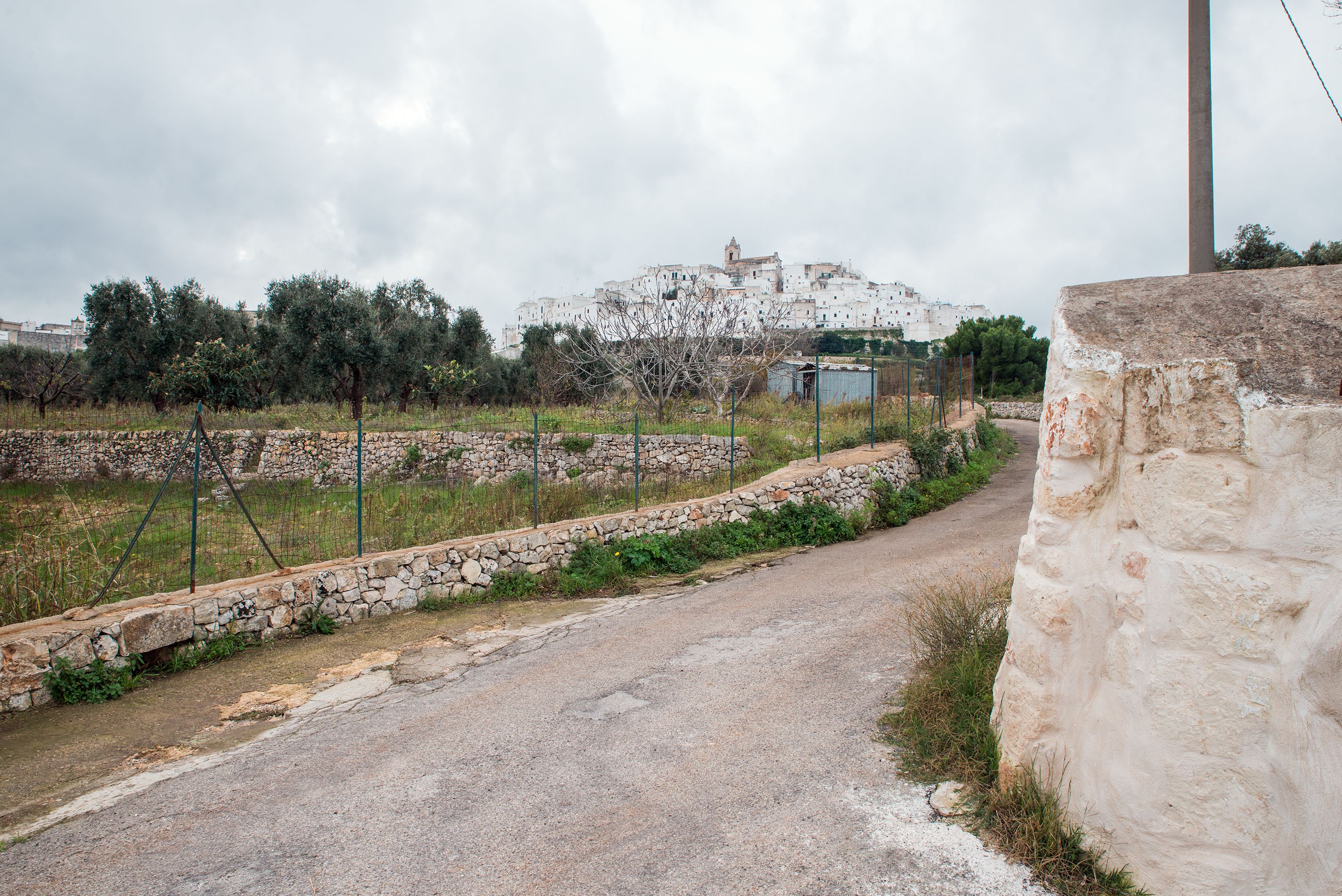 A rural road leading to a white building with a bell tower, surrounded by stone walls and vegetation.