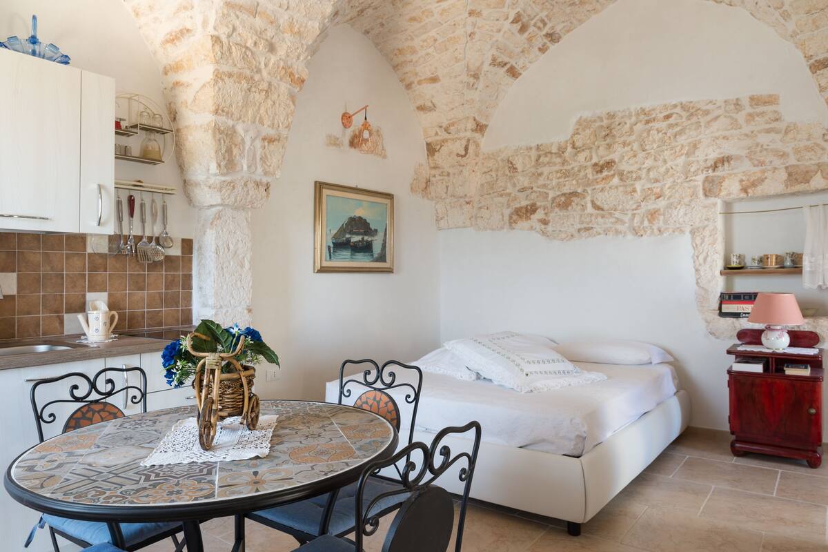 Una habitación acogedora con pared de piedra, un sofá blanco, una mesa de comedor con sillas y una zona de cocina con armarios y fregadero.