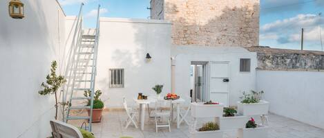 Un patio con paredes blancas, una mesa y sillas blancas, un cojín verde, una escalera y plantas en macetas.