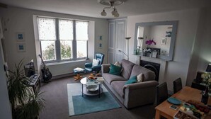 A living room with a sofa, a coffee table, a dining table with food, a chair, a potted plant, and a large window. - Verity and Ilfracombe Harbour View (Ilfracombe)