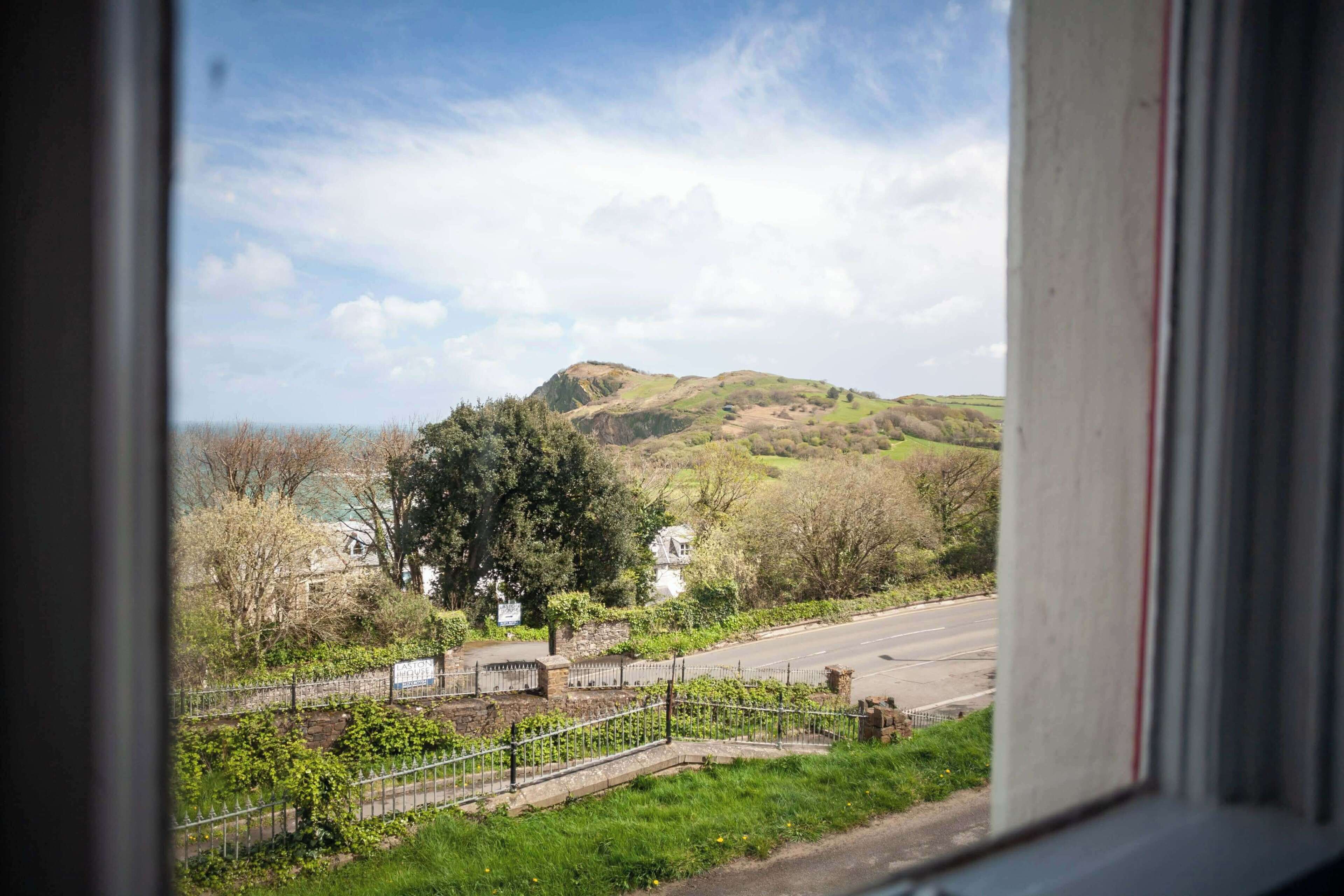 A view through a window of a road leading to a hill with a statue and a stone wall.