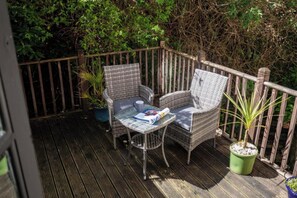 A wooden deck with wicker chairs and a glass table, surrounded by greenery.