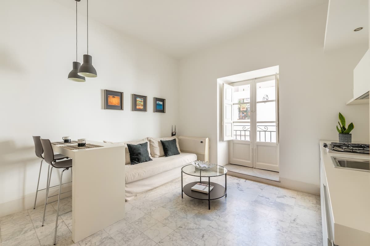 A modern living room with a sofa, two chairs, a glass coffee table, and a kitchen area visible through an open door.