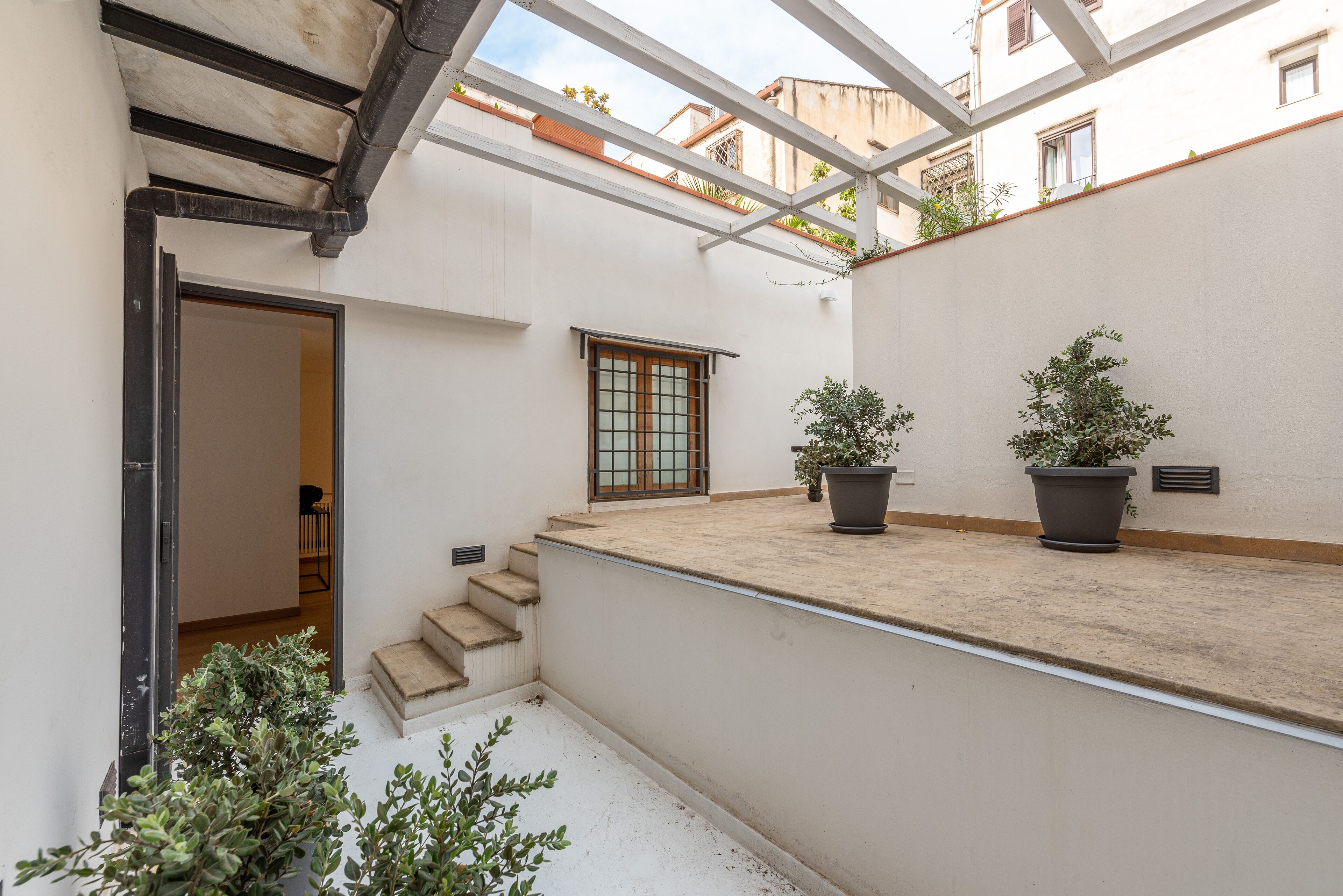 Un patio estrecho con una pequeña escalera hacia una puerta, plantas en macetas y una ventana con celosía de metal.