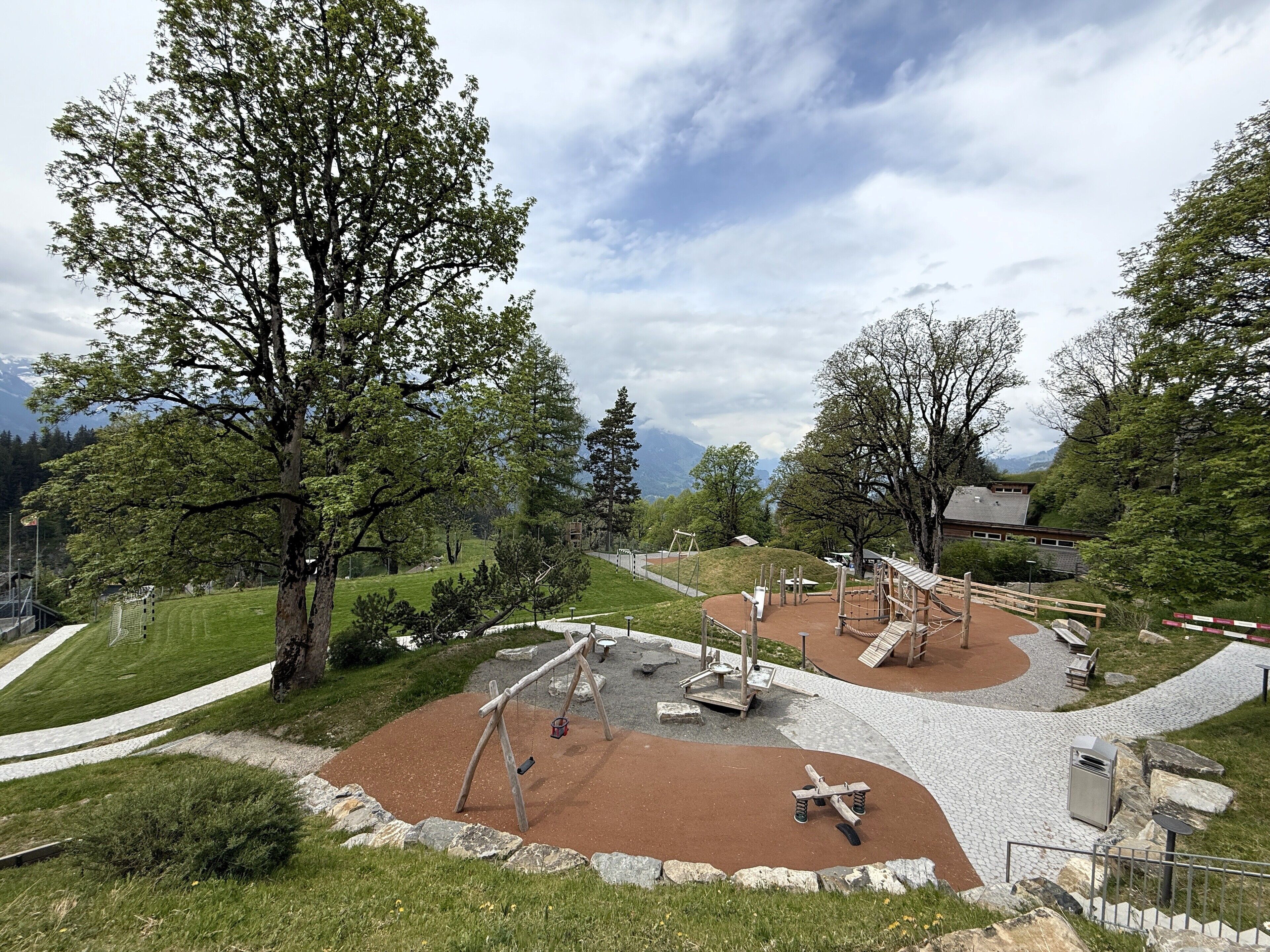 Une aire de jeux avec des balançoires, des structures d’escalade et un bac à sable, le tout entouré d’arbres et offrant une vue sur la montagne.