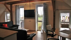 A cozy living room with a black leather sofa, a dining table with chairs, a flat-screen TV mounted on the wall, and large windows with curtains. - Pension Abertham (Abertamy)