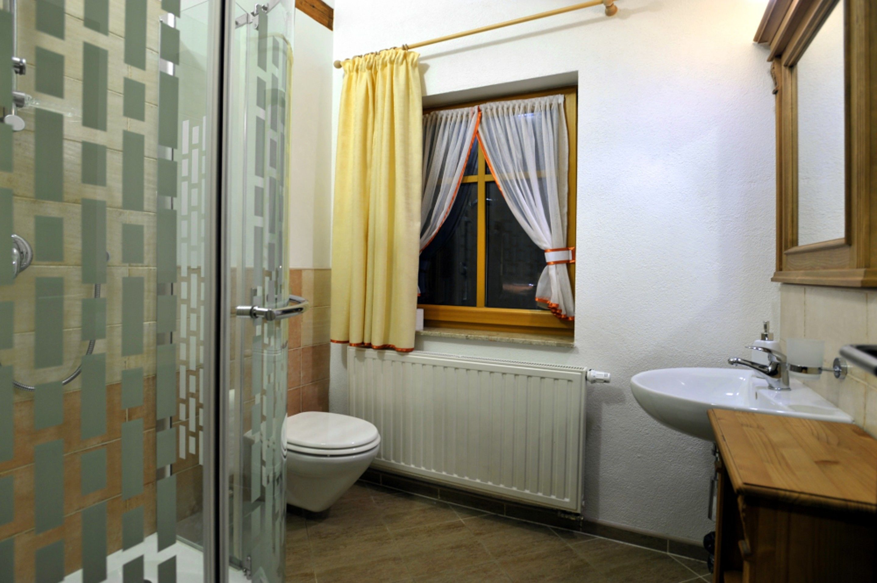 Une salle de bain comprenant des toilettes, un lavabo, une fenêtre avec des rideaux, un espace douche et un meuble en bois.