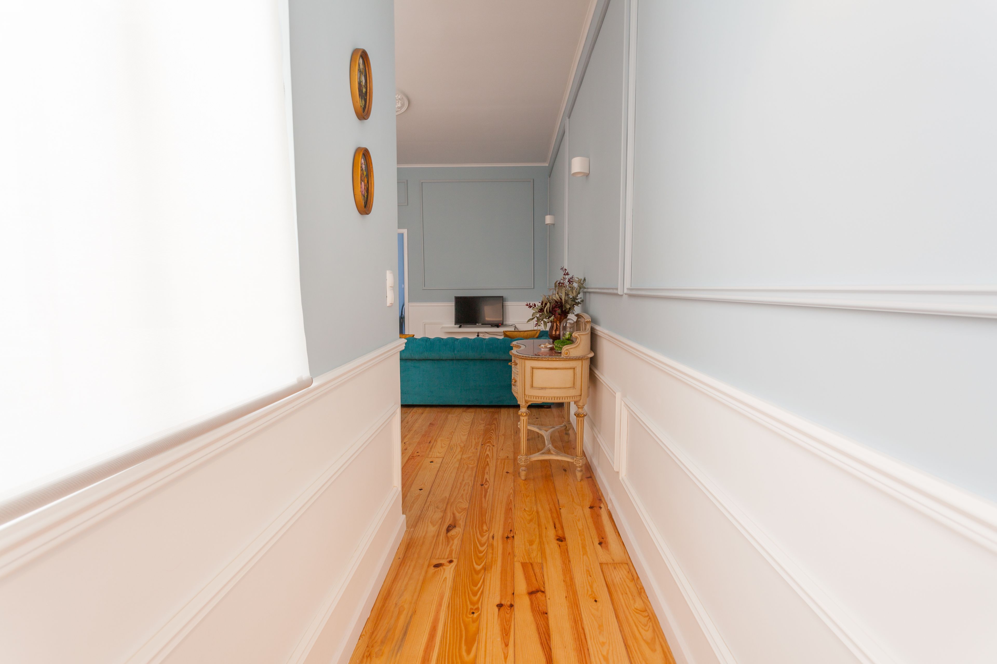 A narrow hallway with wooden flooring, a teal sofa, a small table with a vase, and a television in the background.