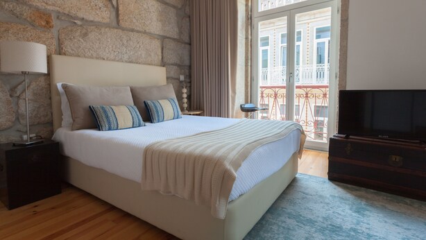 Un dormitorio con pared de piedra, una cama con colcha y almohadas blancas, un televisor sobre un mueble de madera y un balcón con barandilla.