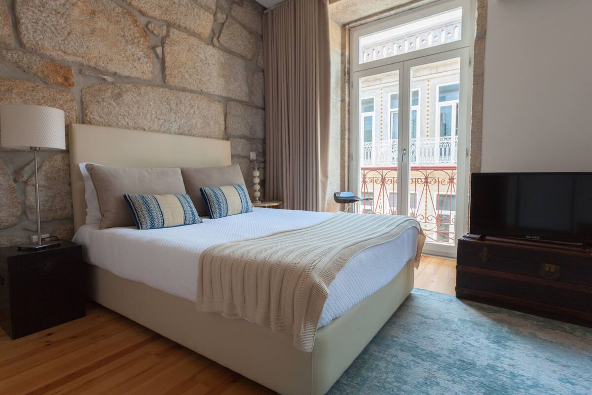 A bedroom with a stone wall, a bed with a white comforter and pillows, a TV on a wooden cabinet, and a balcony with a railing.