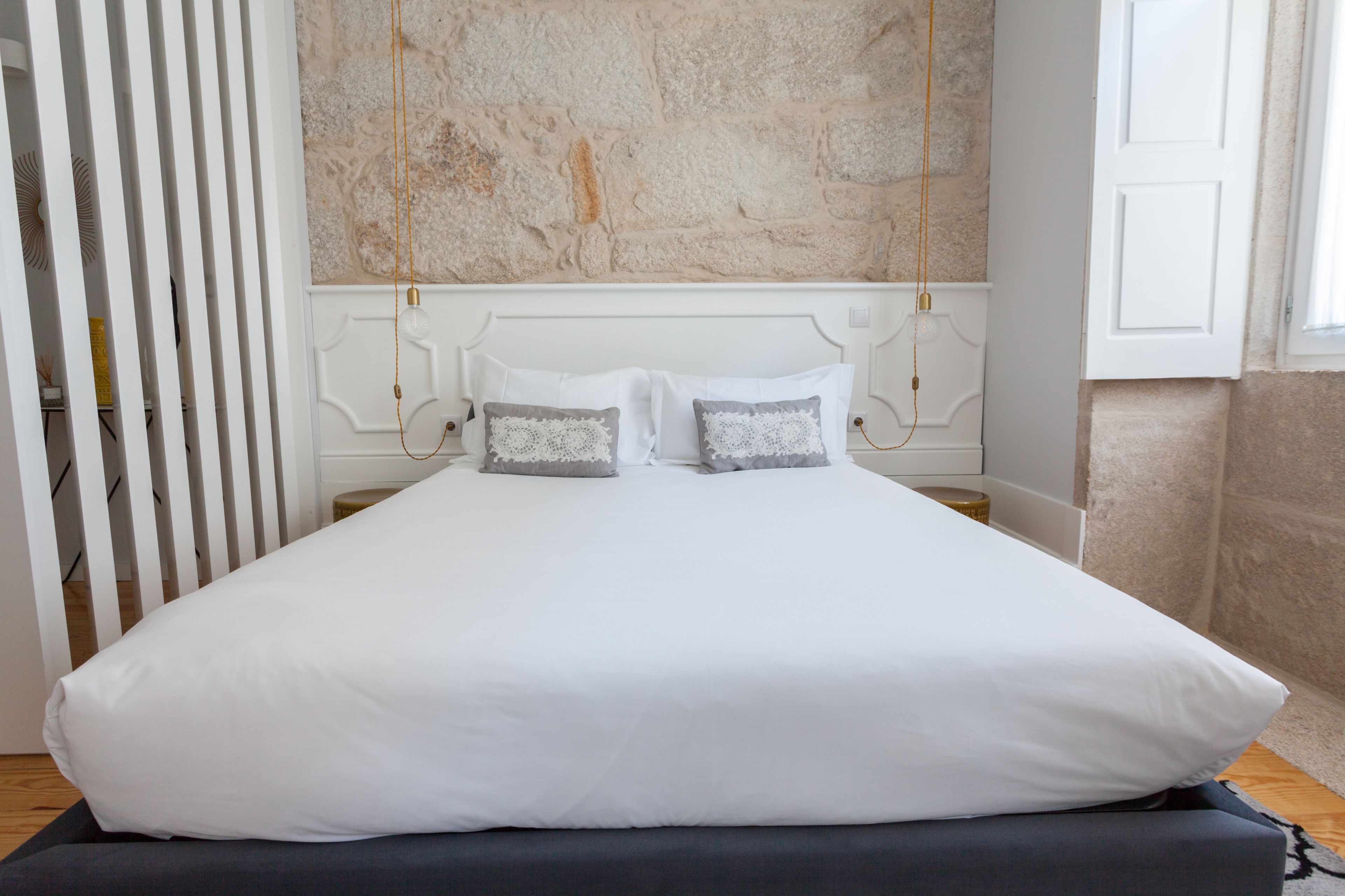 Un letto rifatto con lenzuola bianche e due cuscini decorativi, una parete in pietra sullo sfondo e una porta bianca a destra.