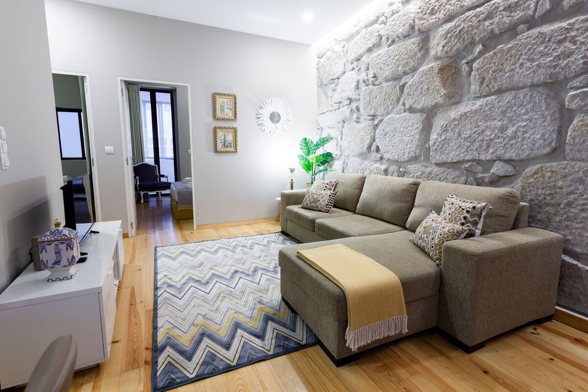 A modern living room with a stone wall, a grey sofa, a patterned rug, and a small table with a decorative item.
