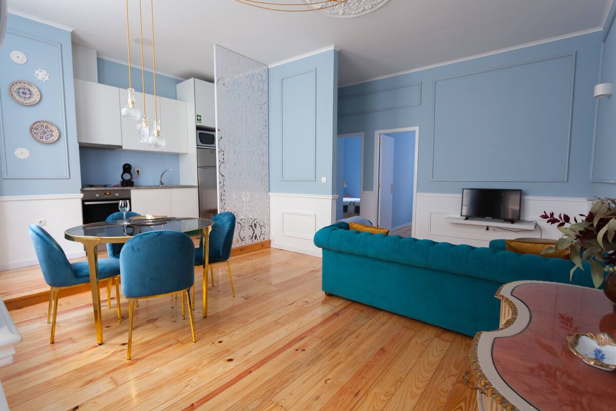 A modern living room with a teal sofa, a dining table with chairs, and a kitchen area with a stove and cabinets.