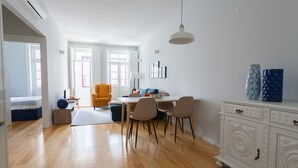 Una sala de estar moderna con comedor, suelo de madera, un sofá, una mesa de comedor con sillas, un armario blanco y un lámpara colgante.