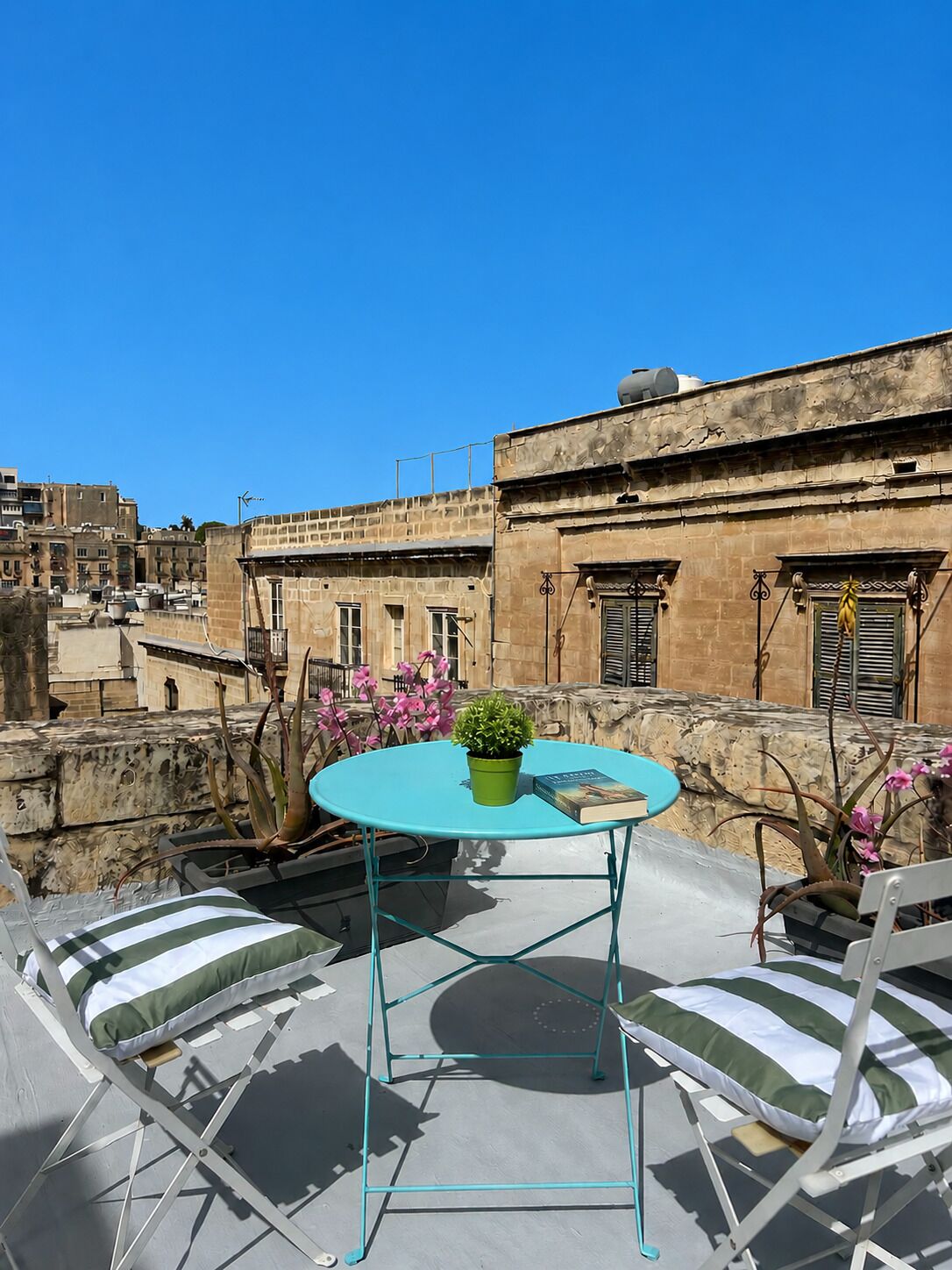 A rooftop terrace with a turquoise table, two striped lounge chairs, a potted plant, and a book.