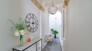 Un pasillo con suelo de mármol, una consola con un jarrón de flores, un reloj, una silla y una lámpara colgante.