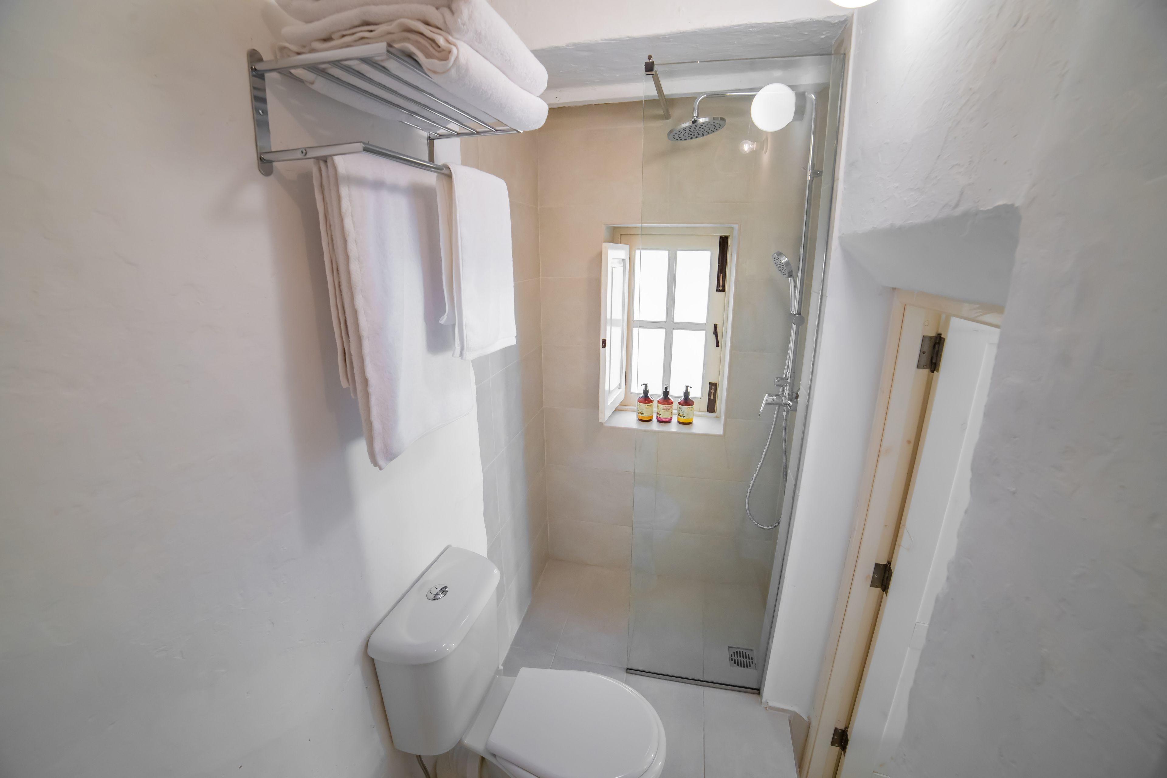 A bathroom with a toilet, a glass shower enclosure, a towel rack, and a window with bottles on the windowsill.