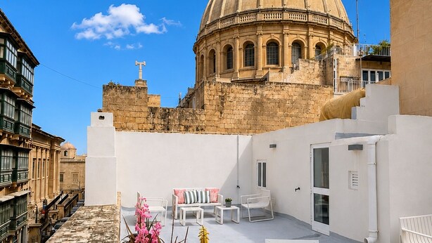 Una terraza en la azotea con vistas a un edificio histórico con cúpula, muebles blancos y un cielo azul despejado.