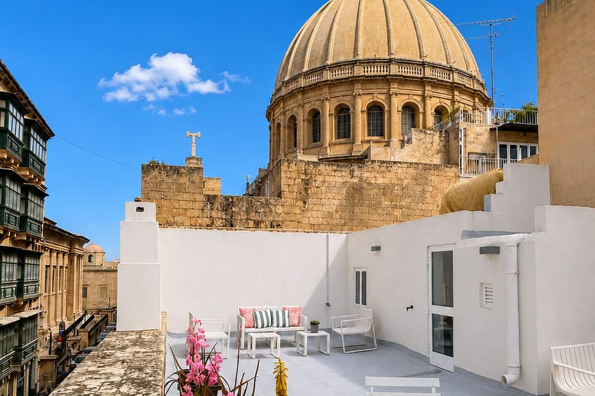 Una terraza en la azotea con vistas a un edificio histórico con cúpula, muebles blancos y un cielo azul despejado.