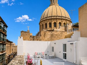 Una terraza en la azotea con vistas a un edificio histórico con cúpula, muebles blancos y un cielo azul despejado.