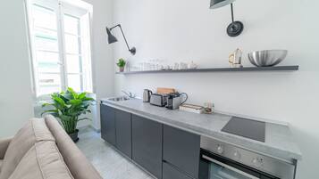 Una cocina moderna con armarios grises, un horno empotrado, una encimera con un hervidor, tostadora y un tazón. Hay una planta en el suelo y una ventana con persianas.