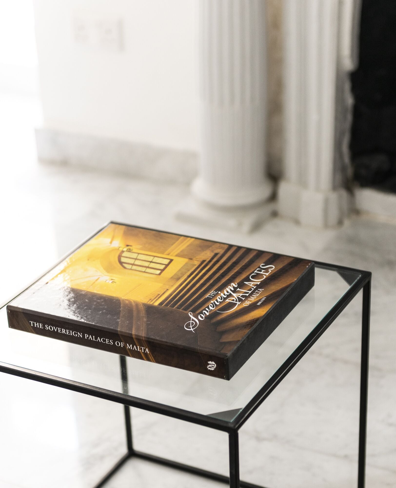 A book titled "The Sovereign Palaces of Malta" on a glass table in a room with white columns.