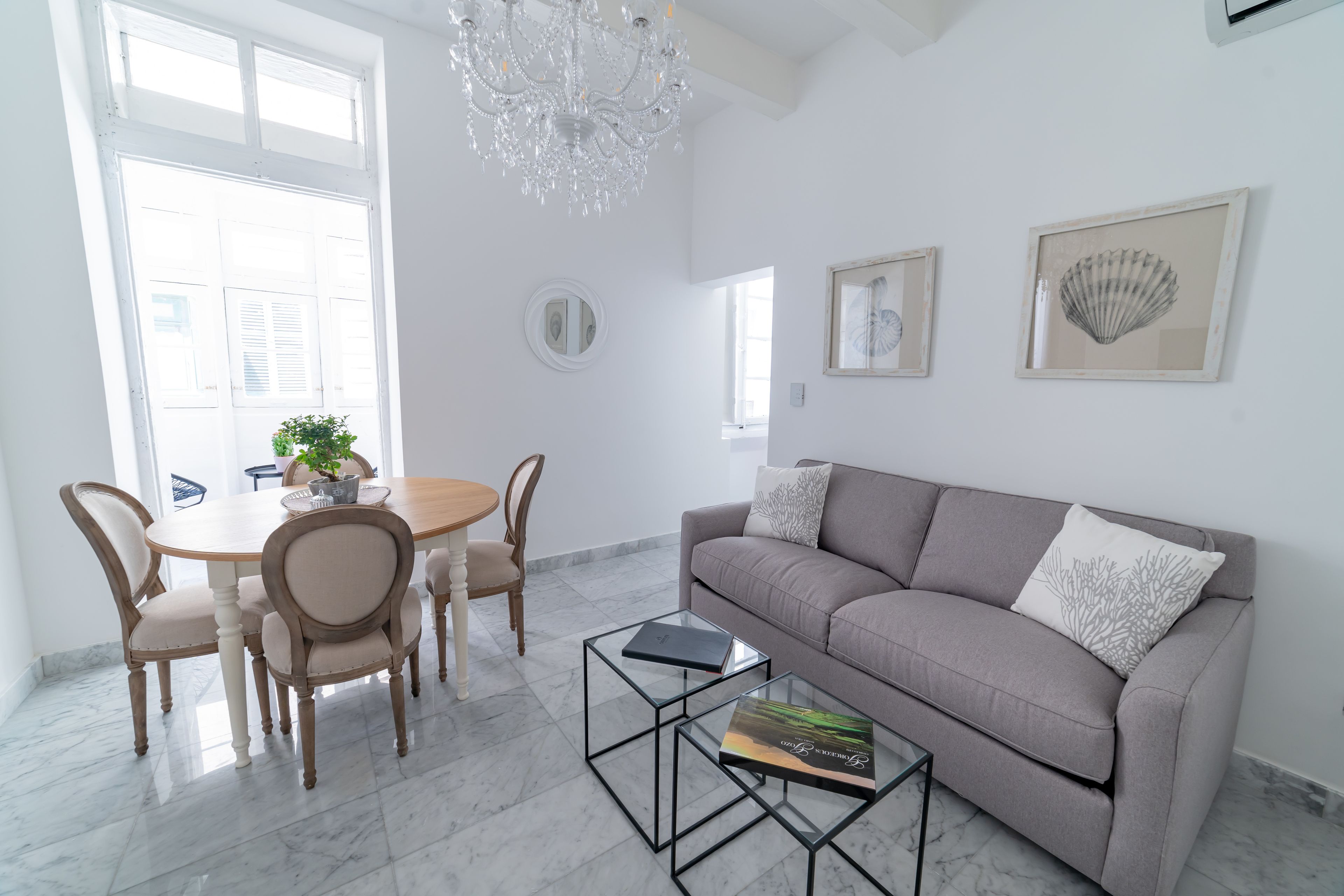 Een moderne woonkamer met een grijze bank, een ronde eettafel met stoelen, een kroonluchter en kunstwerken in lijsten aan de muren.