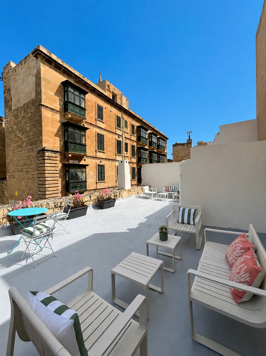 An outdoor seating area with white furniture and potted plants, a building with stone facade and multiple windows, and a clear blue sky.