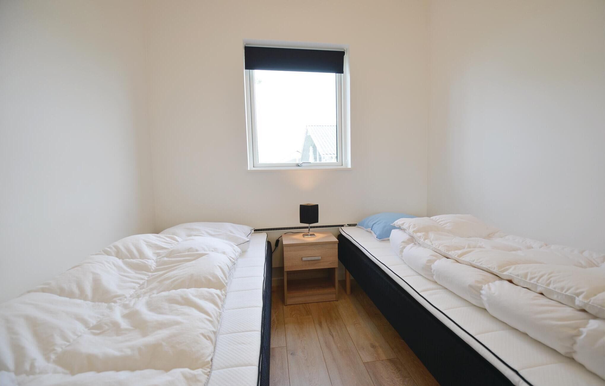 Quarto pequeno com cama de solteiro, janela, mesa de cabeceira e abajur, além de um travesseiro azul.