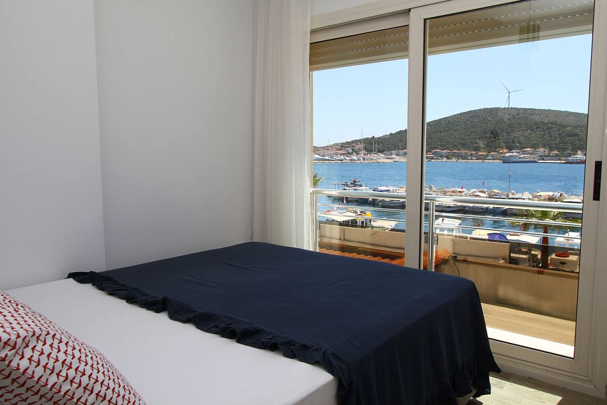 Quarto com cama, varanda com vista para o mar e barcos, e uma turbina eólica ao longe.