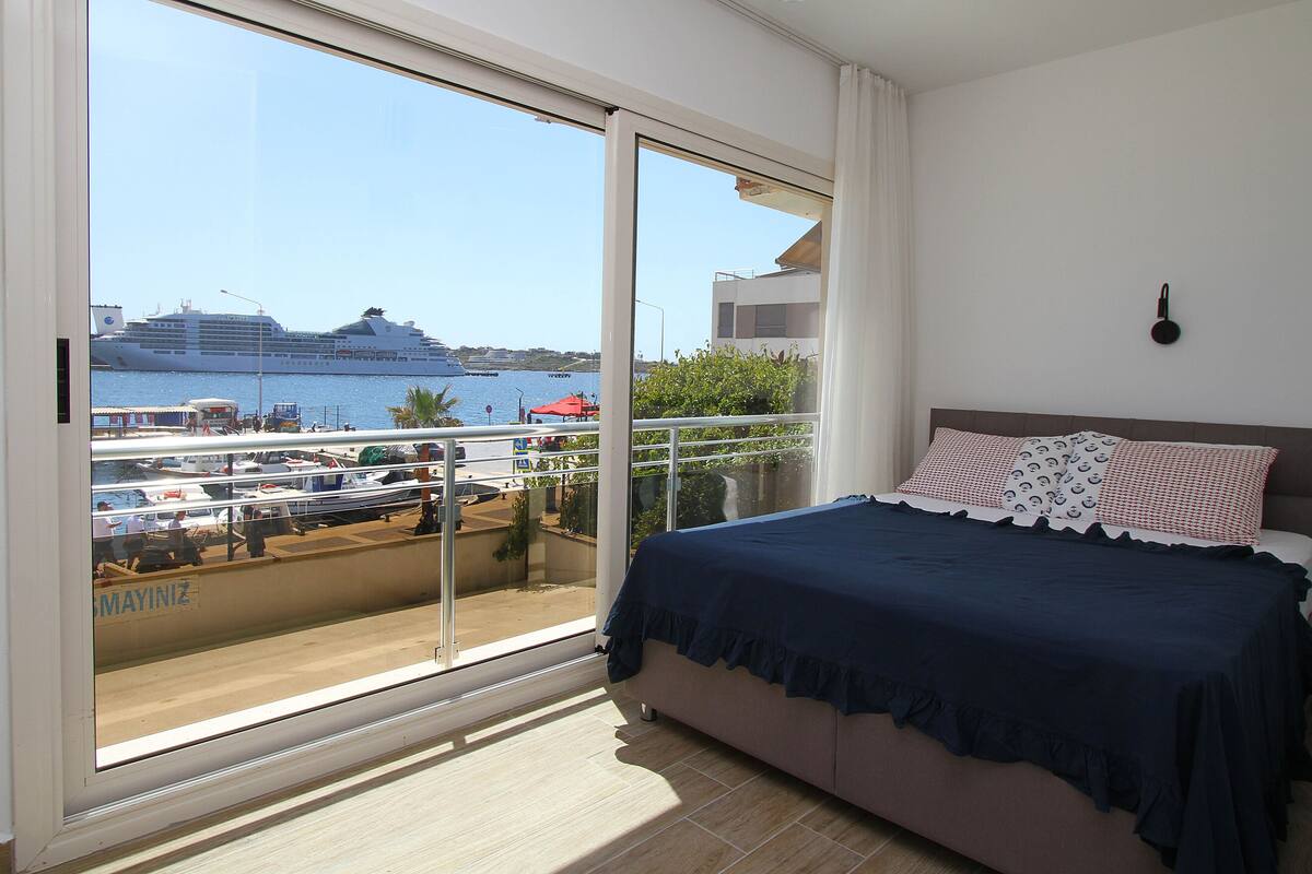Quarto com uma grande porta de vidro deslizante que dá para uma varanda, uma cama com colcha azul escura, e vista de um navio de cruzeiro atracado em um calhave.