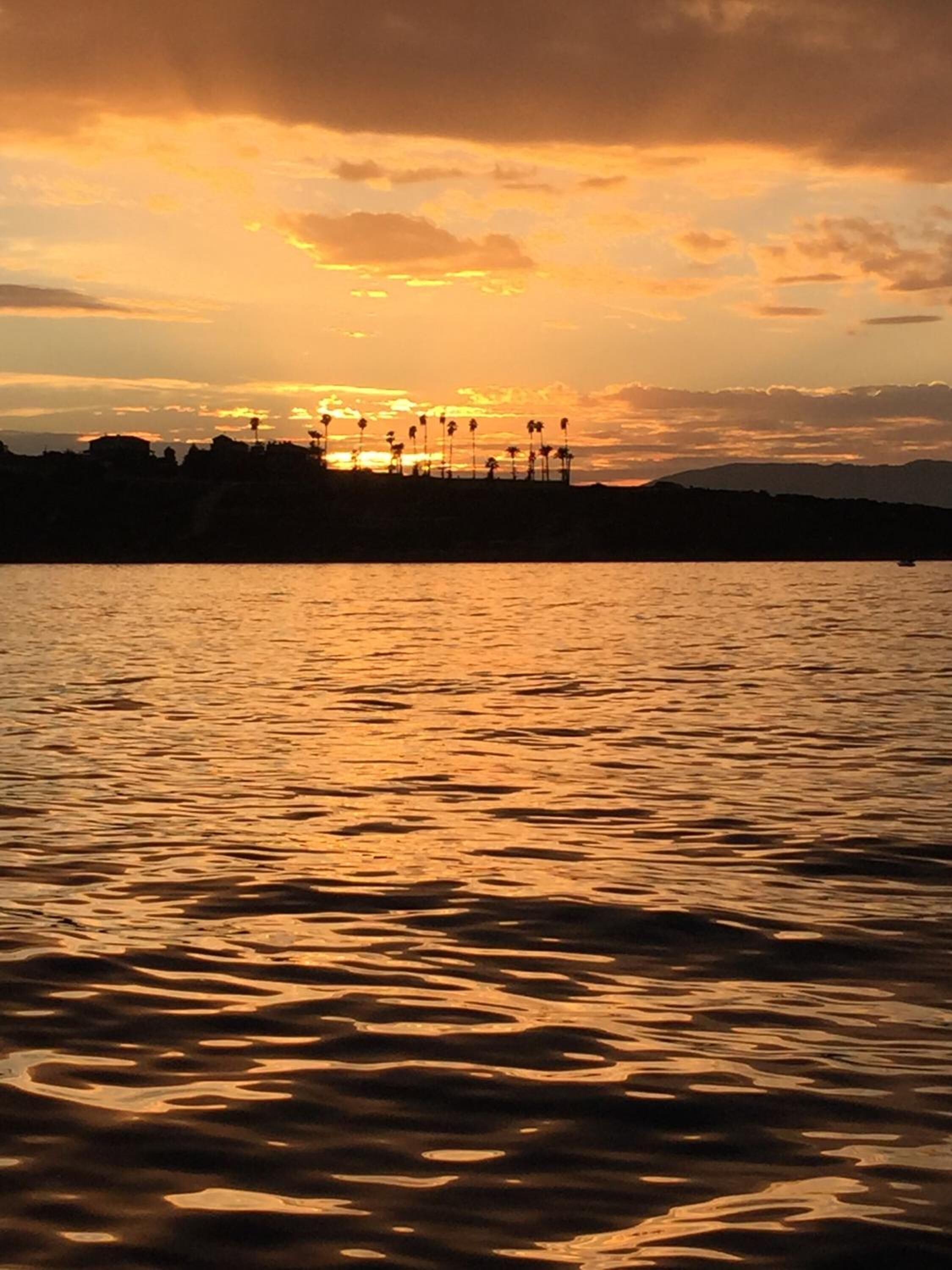 A sunset over a body of water with silhouettes of palm trees and a distant shoreline.