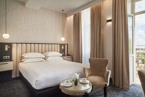 Habitación de hotel con una cama grande, una silla, una mesa con tetera y tazas, y vistas al exterior.