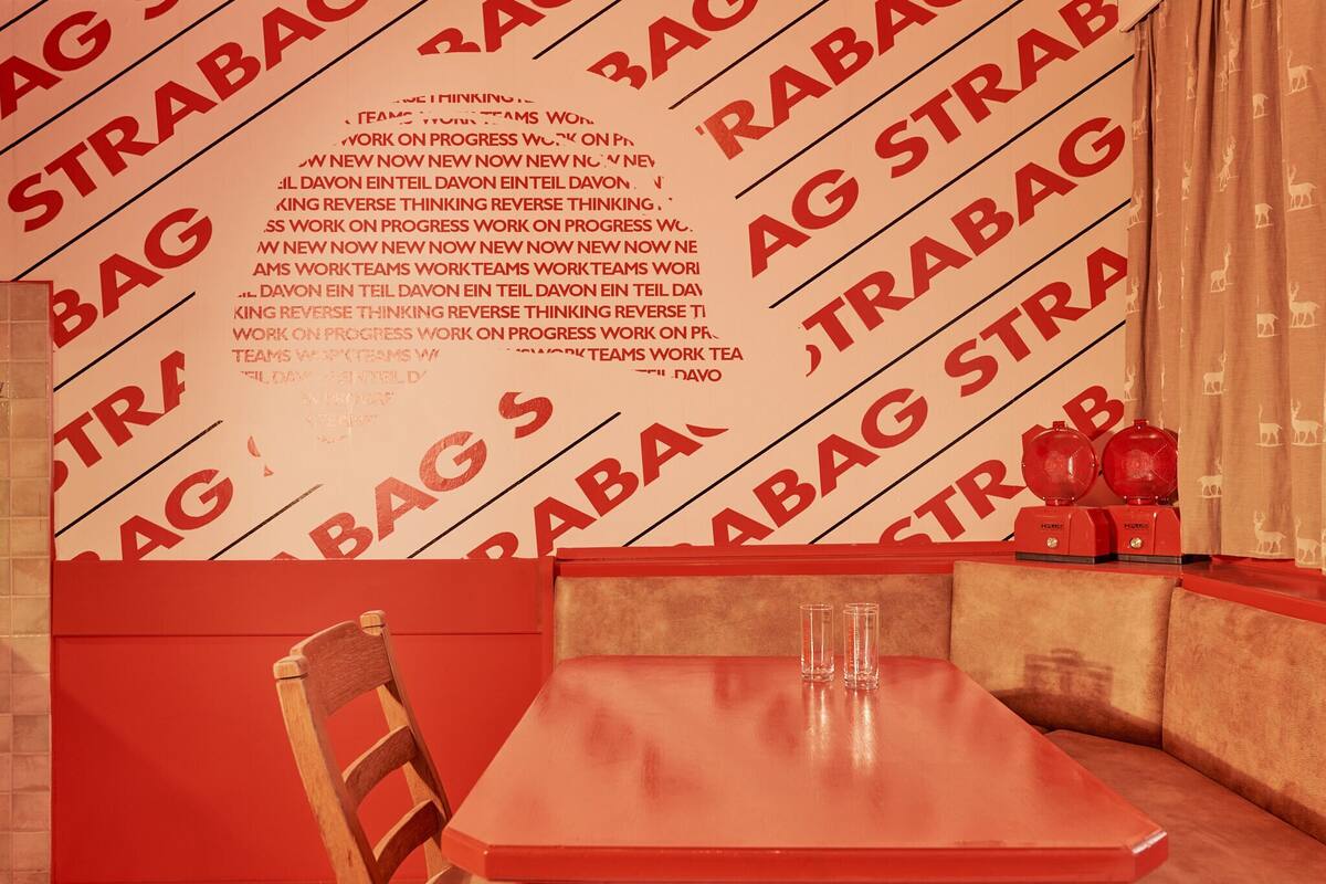 A booth with a red table and a wooden chair, a wall with 'RABAG STRABAG' text, and two red objects on the table.