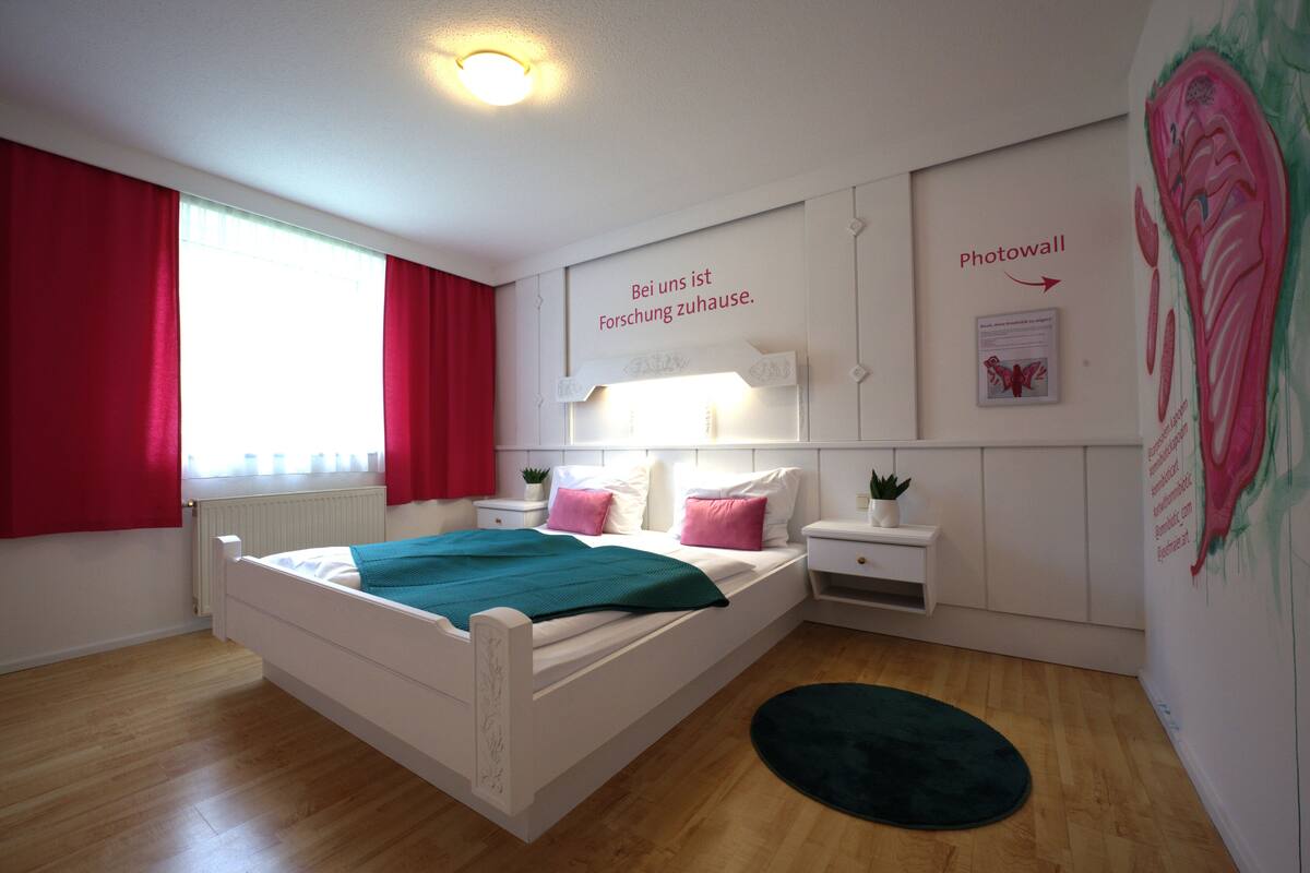 A bedroom with a bed, a nightstand, a wall-mounted lamp, a window with red curtains, and a wall with a 'Photowall' sign.