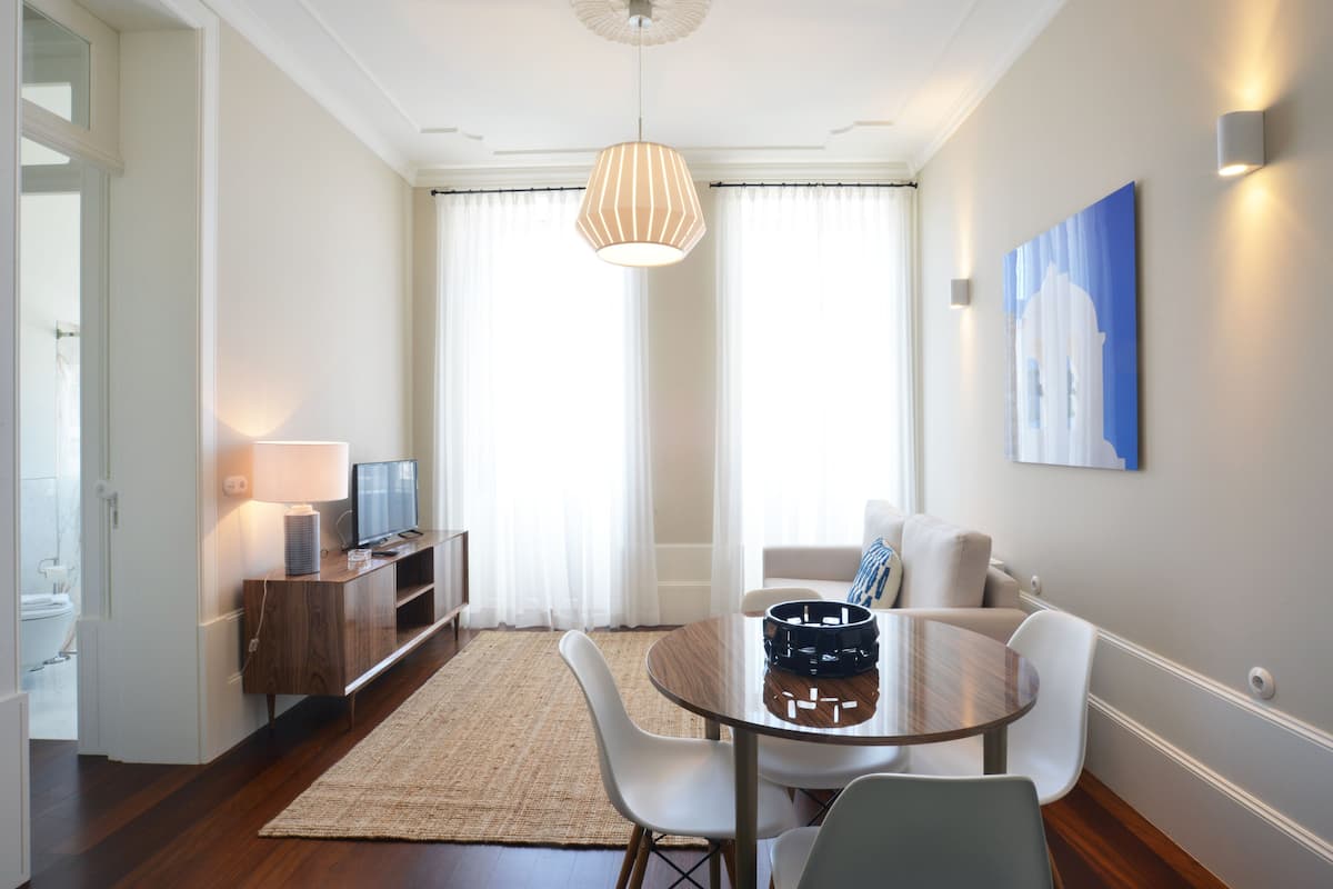 A modern living room with a round dining table, white chairs, a wooden cabinet, a television, and a hanging light fixture.