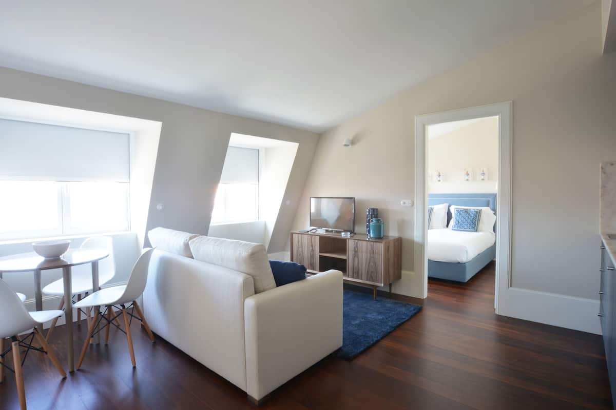 A modern living room with a sofa, dining table, and chairs. There is a TV on a wooden cabinet and a bed with a blue headboard in the background.