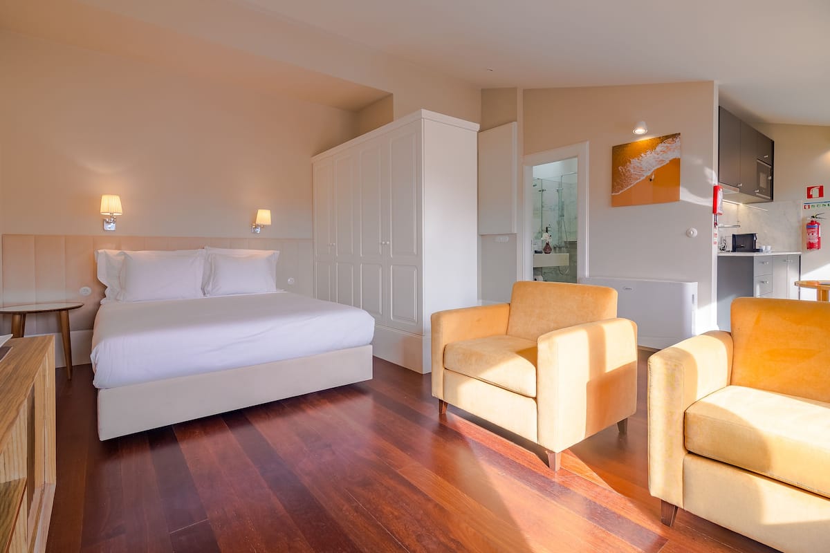 A hotel room with a bed, two armchairs, a small table, a wardrobe, a TV, and a kitchen area.