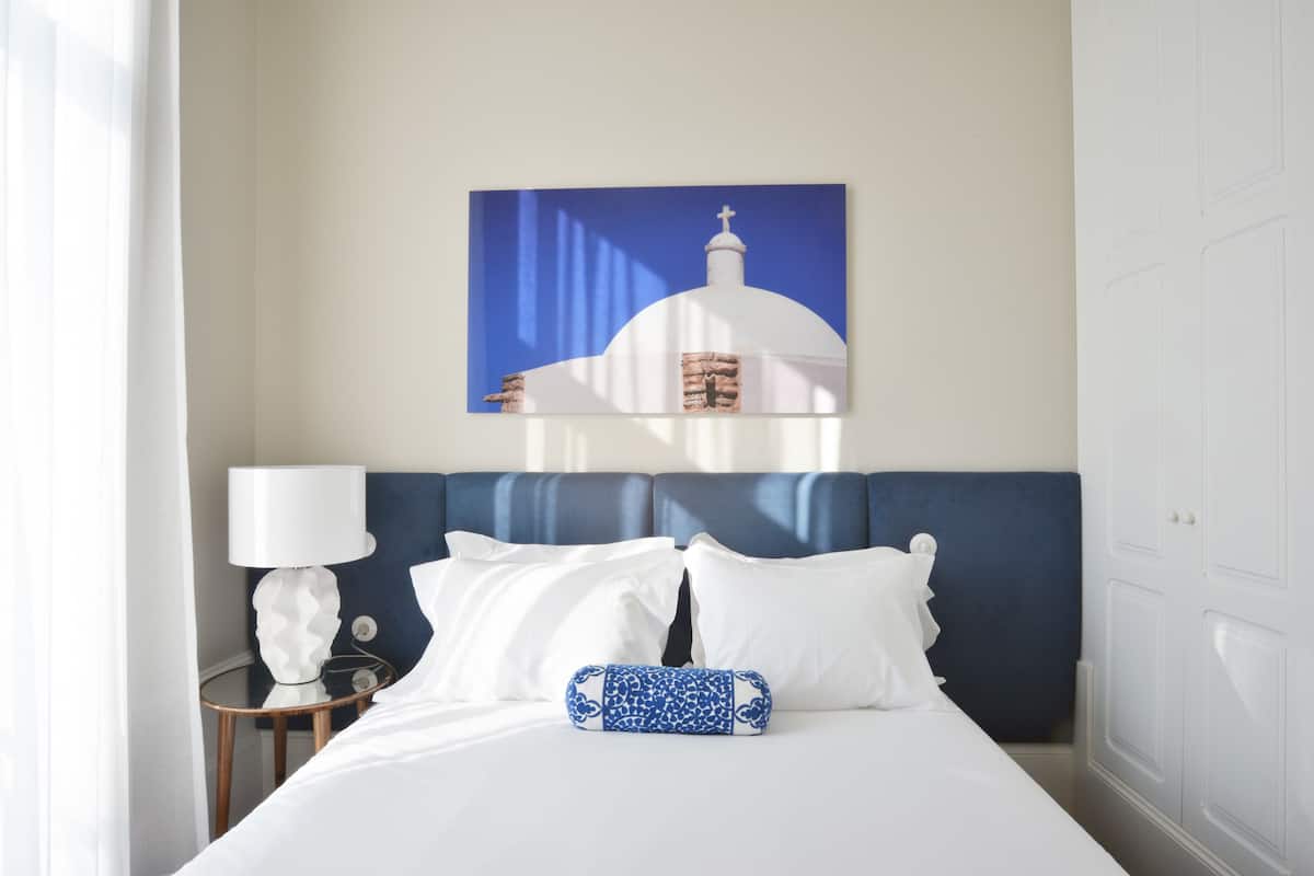 A hotel room with a bed, white bedding, a blue headboard, a white lamp, and a painting of a white building with a cross on top.