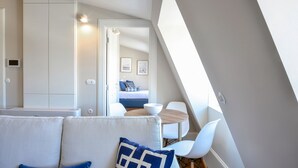 A modern, well-lit room with a sofa, a dining table, and a view of a bedroom with a bed and a wall-mounted light fixture. - YOUROPO - Market (Porto)