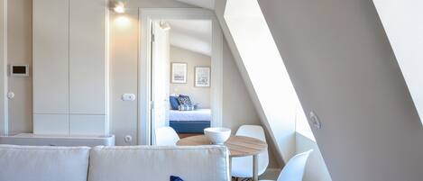 A modern, well-lit room with a sofa, a dining table, and a view of a bedroom with a bed and a wall-mounted light fixture.