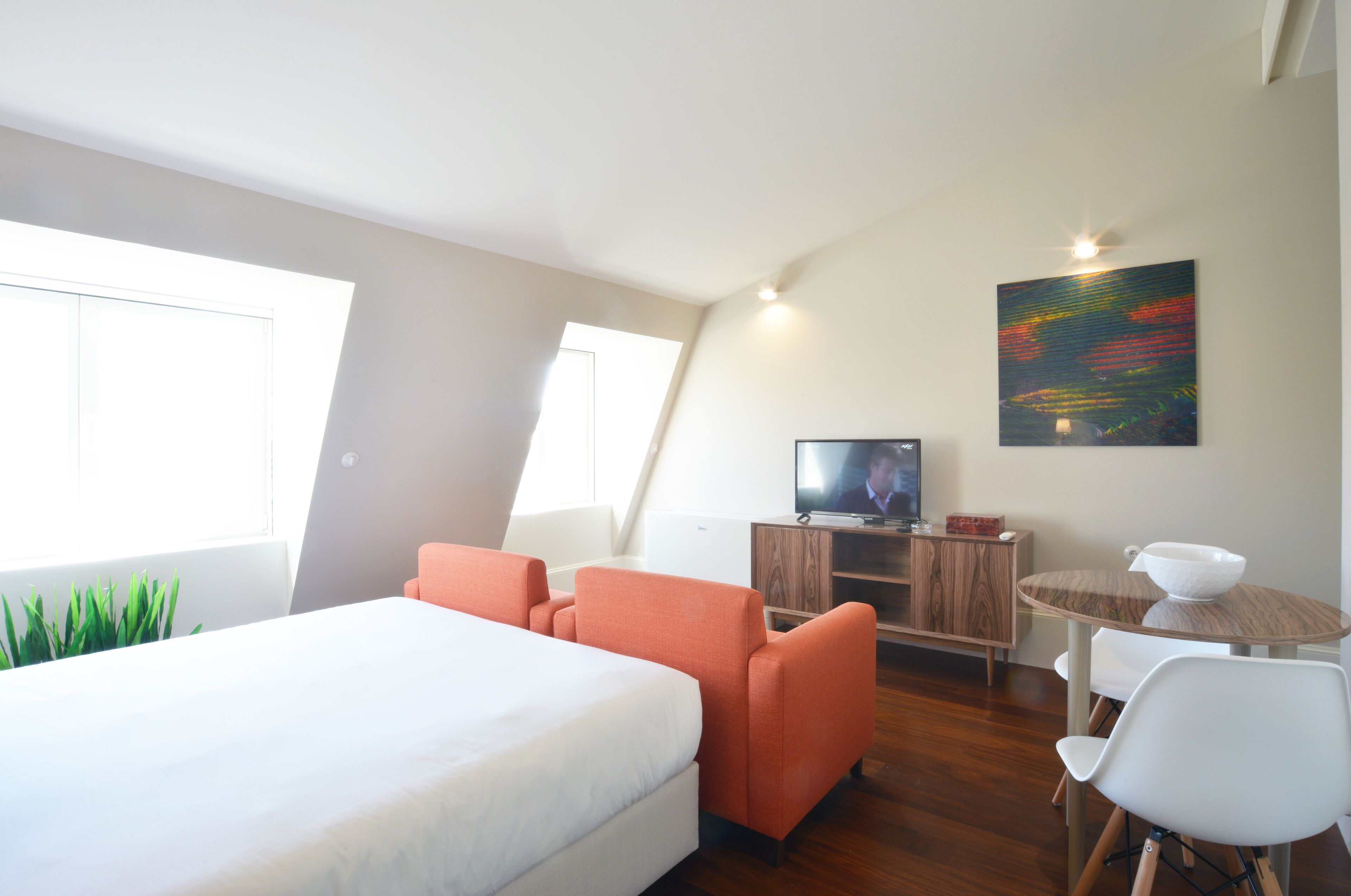 A modern hotel room with a bed, orange armchairs, a dining table with chairs, a TV, and a painting on the wall.