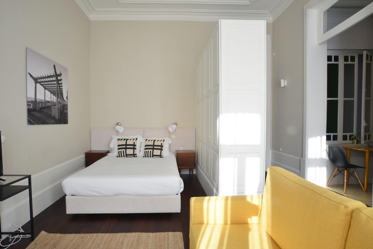 A bedroom with a bed, a yellow sofa, a nightstand, a lamp, and a painting on the wall.