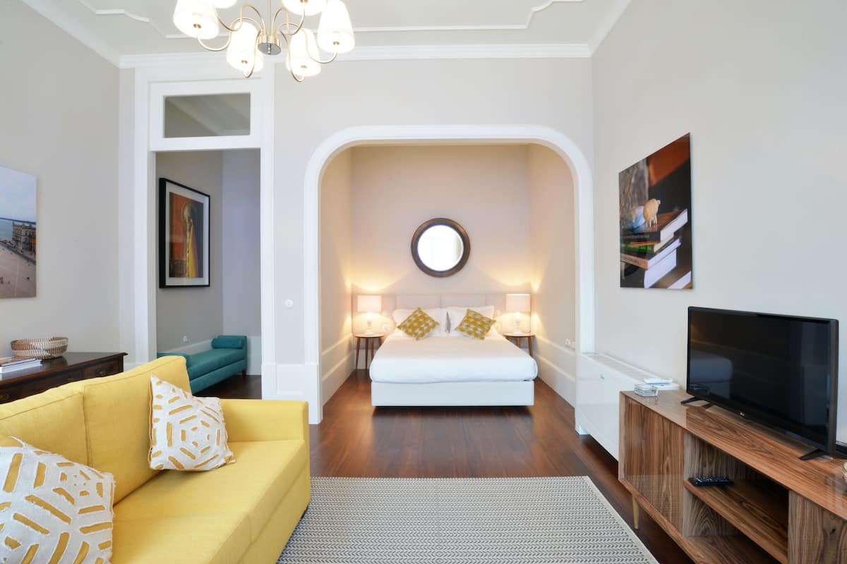 A modern living room with a yellow sofa, a white bed, and a television on a wooden stand.