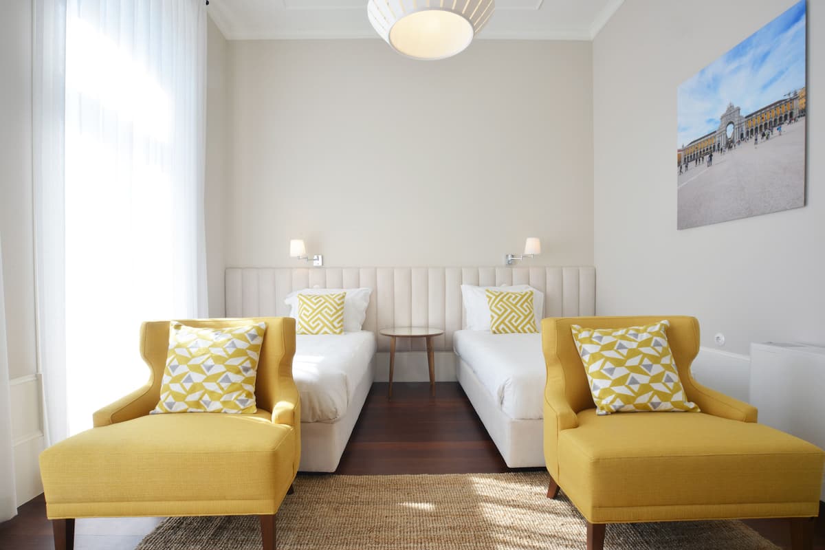A hotel room with two single beds, a small round table, and two yellow armchairs.