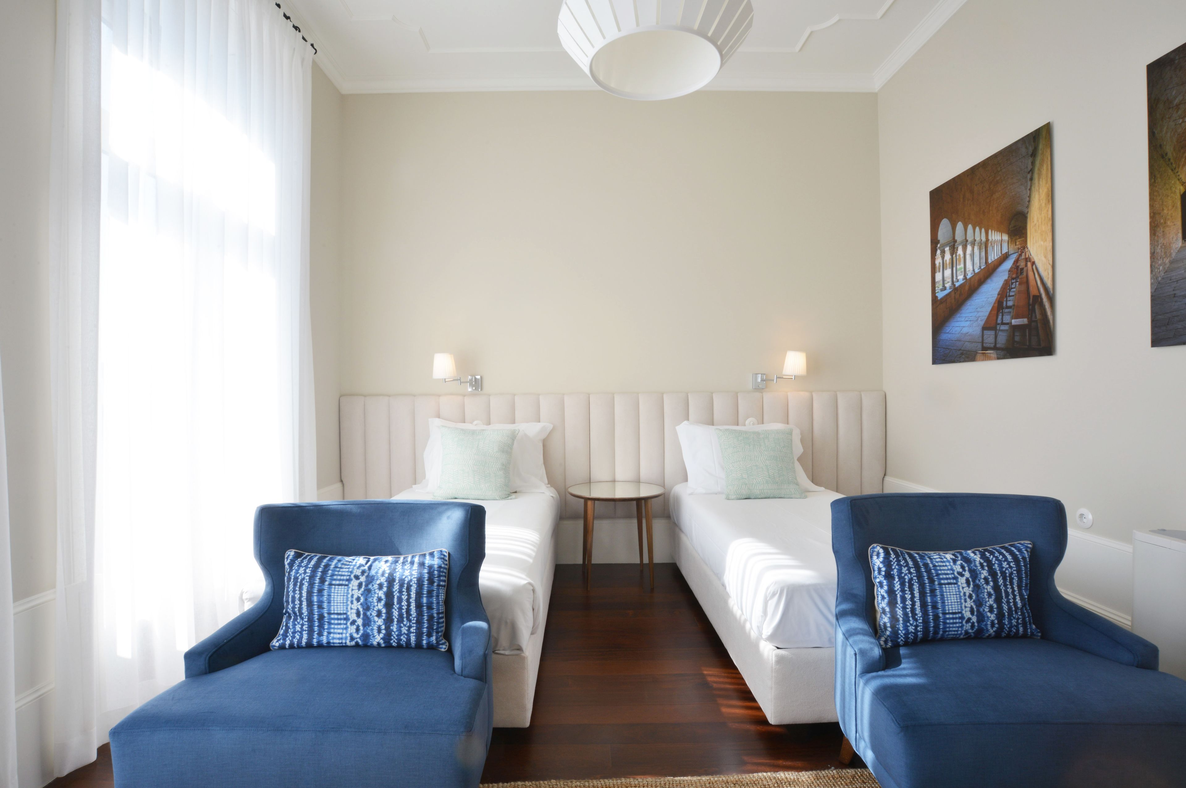 A hotel room with two single beds, a small round table, and two blue armchairs.