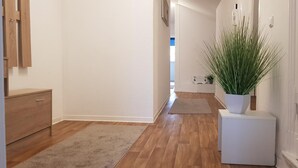 Un couloir avec un plancher en bois, une porte blanche, un petit meuble blanc et une plante en pot.
