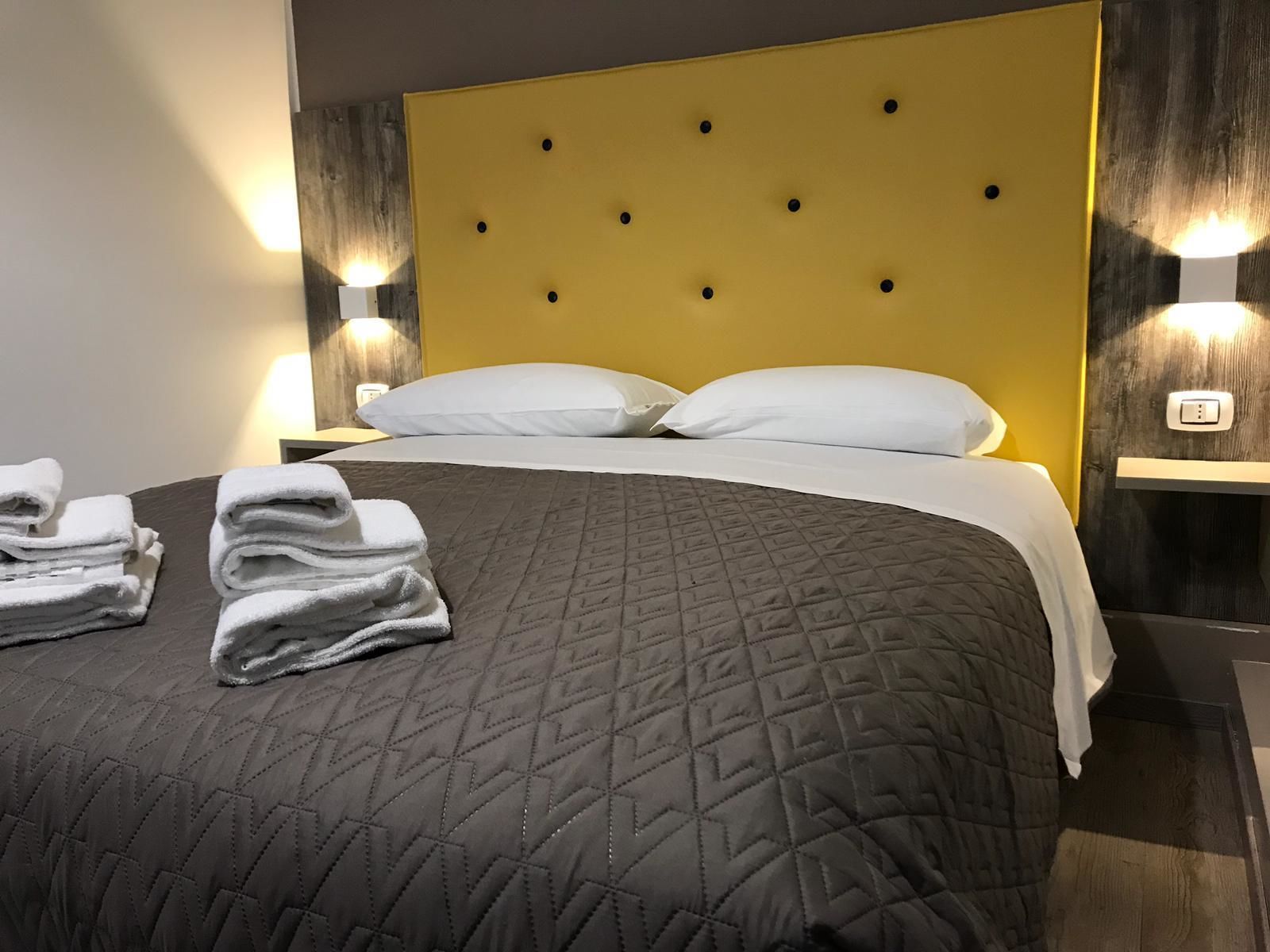 A neatly made bed with a quilted gray bedspread and white pillows, two white towels on the bed, and two bedside lamps on wooden nightstands.